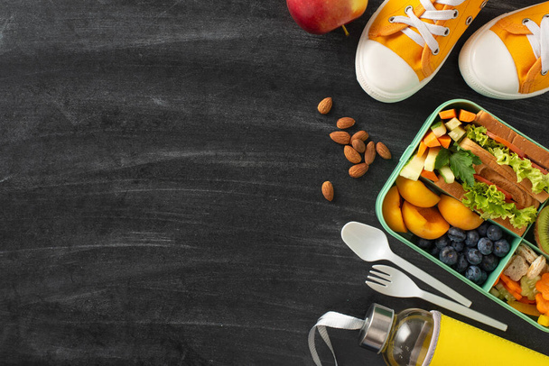 School snack ensemble promoting health. Top view revealing lunchbox filled with fruits, vegetable sandwich, berries, nuts, water bottle, cutlery, shoes on blackboard surface, space for text or ad - Photo, Image