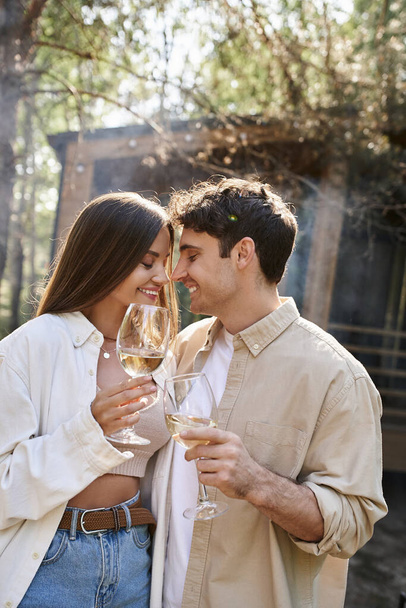 Smiling brunette couple holding wine and standing with closed eyes near vacation house outdoors - Foto, Imagem