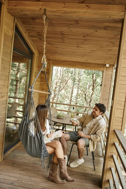 Θετικό ρομαντικό ζευγάρι με κρασί και φαγητό που μιλάει και ξοδεύει χρόνο στη βεράντα του θερινού σπιτιού - Φωτογραφία, εικόνα