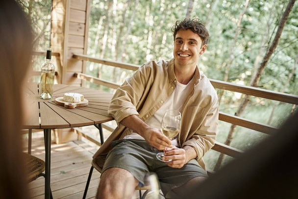 Smiling brunette man holding wine near cheese and blurred girlfriend on terrace of vacation house - Photo, Image