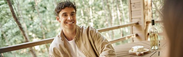Улыбающийся брюнетка мужчина смотрит на подругу возле вина и сыра на террасе дома отдыха, баннер - Фото, изображение