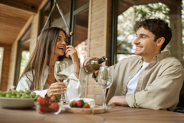 Mujer sonriente comiendo fresa cerca de novio con vino en la terraza de la casa de vacaciones de madera - Foto, Imagen