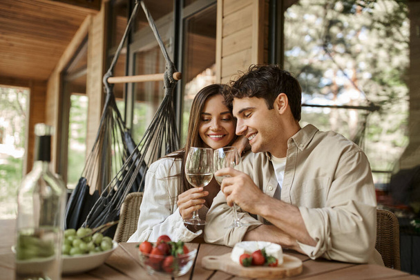 Smiling romantic couple clinking with wine near blurred food on terrace of wooden vacation house - Φωτογραφία, εικόνα