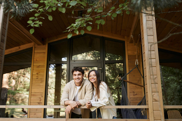 Χαμογελαστό ρομαντικό ζευγάρι κοιτάζοντας κάμερα, ενώ στέκεται στη βεράντα του ξύλινου εξοχικού σε εξωτερικούς χώρους - Φωτογραφία, εικόνα