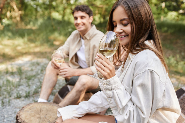 Sonriente joven mujer sosteniendo un vaso de vino y sentado cerca borrosa novio al aire libre - Foto, Imagen