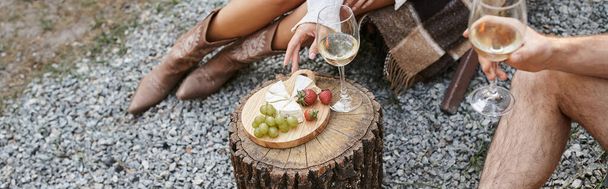 Vista recortada de la pareja sosteniendo vino cerca de frutas frescas y queso al aire libre durante el picnic, pancarta - Foto, Imagen