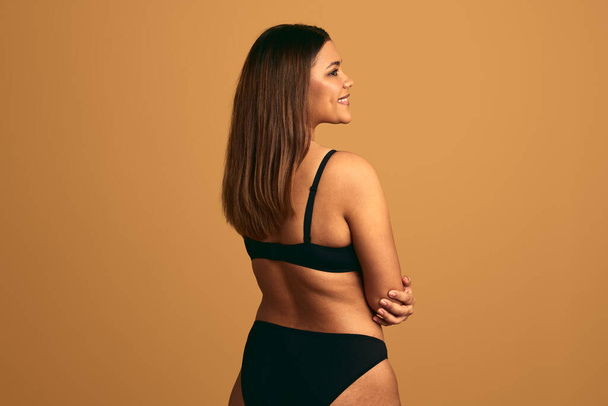 Back view of positive young plus sized female model in black lingerie with long hair and arms crossed, looking away against beige background in studio - Foto, Imagen