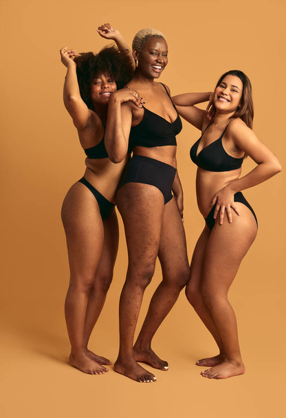 Full body of happy young multiracial female models in black lingerie smiling and looking at camera while standing against yellow background together in studio - Foto, Imagen