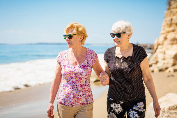 Dos ancianas están caminando por la costa rocosa, hablando y riendo, pasando un rato agradable juntas. - Foto, Imagen