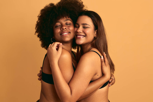 Positive young multiracial female friends in underwear embracing and smiling while standing against beige background with closed eyes in studio - Фото, изображение