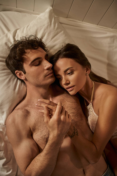 Bovenaanzicht van sexy vrouw in lingerie aanraken hand van shirtloos vriendje op bed in vakantiehuis - Foto, afbeelding