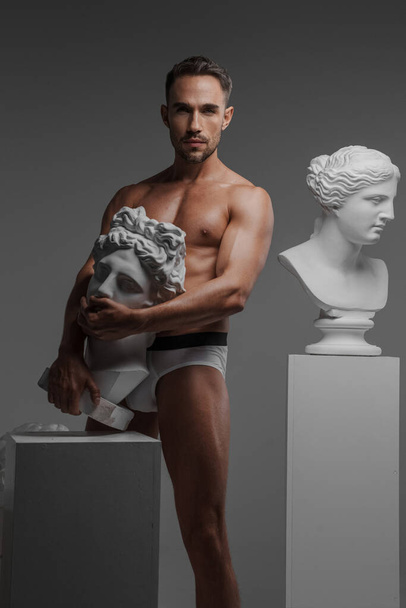Харизматичный мужчина-модель со скульптурным телосложением позирует рядом с бюстом древнегреческой скульптуры - Фото, изображение
