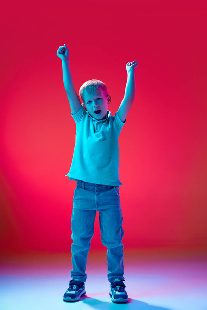 Full-length portrait of cheerful boy, child in casual clothes standing with hands up against pink studio background in neon light. Concept of childhood, lifestyle, emotions, education, fashion, ad - Foto, Imagem