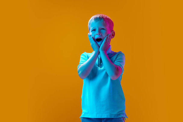 Good surprise. Happy, excited boy, child standing against yellow studio background in neon light. Celebration. Concept of childhood, lifestyle and emotions, education, fashion, care, ad - Foto, Imagem