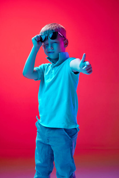 Super good. Portrait of little boy, child in casual clothes and sunglasses against pink studio background in neon light. Concept of childhood, lifestyle, emotions, education, fashion, care, ad - Photo, Image