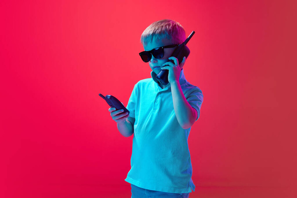 Little boy, child talking on retro, old mobile phone against pink studio background in neon light. Serious look. Concept of childhood, lifestyle, emotions, education, fashion, care, ad - Foto, imagen