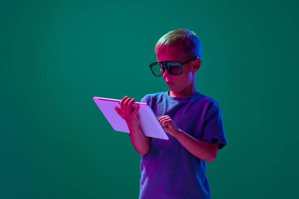 Online games. Little boy, child in sunglasses playing on tablet with excitement against cyan studio background in neon light. Concept of childhood, lifestyle, emotions, education, fashion, care, ad - Photo, Image