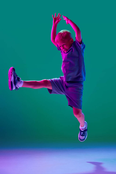 Little boy, child in casual clothes cheerfully jumping, playing against cyan studio background in neon light. Fantasy. Concept of childhood, lifestyle, emotions, education, fashion, care, ad - Photo, image
