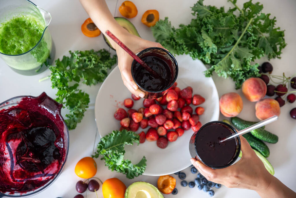 Мама и ребенок держат в руках стаканы с ягодными смузи на фоне кухонного стола с овощами и фруктами. - Фото, изображение