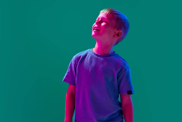 Portrait of little boy, child in casual clothes standing with smile against cyan studio background in neon light. Concept of childhood, lifestyle, emotions, education, fashion, care, ad - Photo, Image