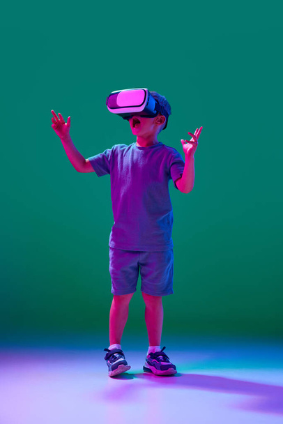 Little boy, child wearing vr glasses against cyan studio background in neon light. Online games, movies. Concept of childhood, lifestyle, emotions, education, modern technologies, ad - 写真・画像