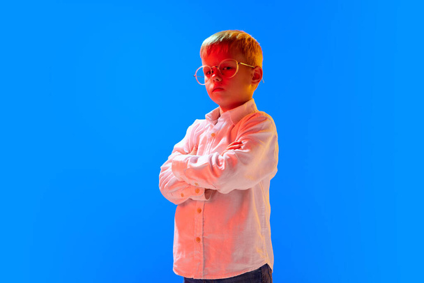 Portrait of smart, serious boy, child, pupil standing in white shirt and glasses against blue studio background in neon light. Concept of childhood, lifestyle, emotions, education, fashion, care, ad - Foto, imagen