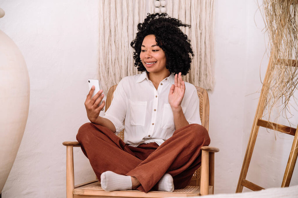 smiling afro american woman using smartphone for video call while sitting on armchair in living room. young female looking at cell phone screen and wave hand starting conversation in online app - Foto, Imagen