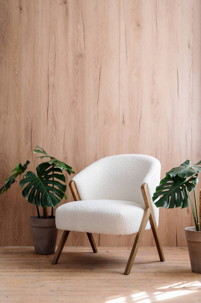 Vertical shot of white comfortable chair in room with wooden planks on wall. Green monstera, houseplants in pots close to soft furniture in lobby. Nordic style interior with hardwood floor. Copy space - Fotografie, Obrázek
