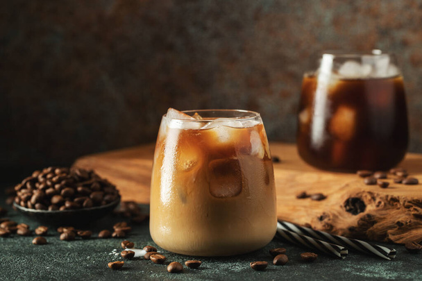 Ice coffee in a tall glass with cream poured over, ice cubes and beans on a dark concrete table. Cold summer drink with tubes on a black background with copy space. - Zdjęcie, obraz