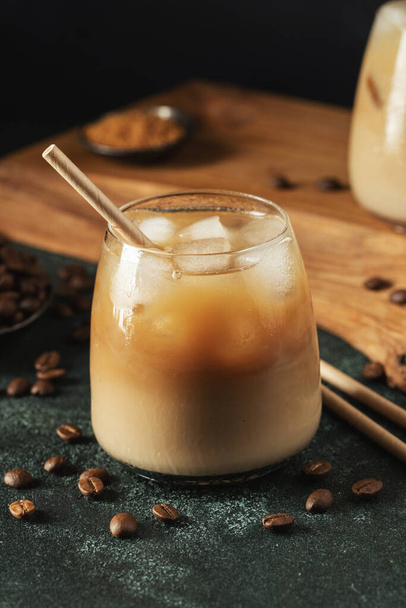 クリームをかけた背の高いガラスの中のアイスコーヒーは、暗いコンクリートテーブルの上に氷のキューブと豆を注ぎました。コピースペースと黒の背景にチューブで冷たい夏の飲み物. - 写真・画像