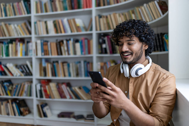 Молодой латиноамериканец улыбается и счастлив, используя онлайн-приложение для обучения, улыбается мужчина в наушниках, сидя в академической библиотеке среди книг. - Фото, изображение
