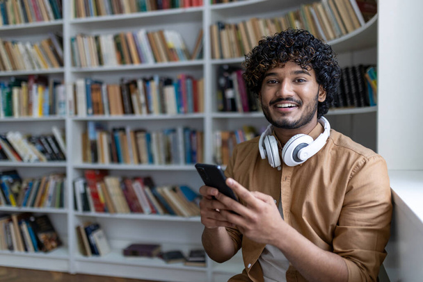 Young hispanic student inside public library studying, smiling and looking at camera, man holding phone, using study app, with headphones among shelves. - Fotó, kép