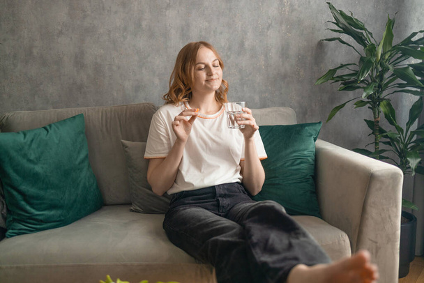 Jeune femme blonde prenant pilule boire de l'eau assis sur le canapé à la maison. Photo de haute qualité - Photo, image