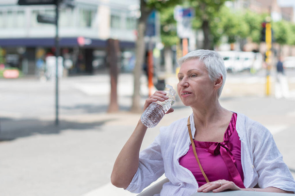 une femme caucasienne de soixante ans avec Bob coupé dans des vêtements d'été élégants boire de l'eau de la bouteille en plastique à la table de café dans la ville, photo de haute qualité - Photo, image