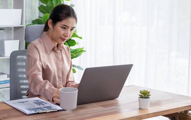 Young asian enthusiastic businesswoman at modern office desk using laptop to work and write notes. Diligent and attractive office lady working on computer notebook in her office work space. - Фото, зображення