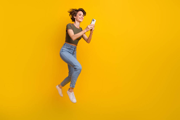 Full length photo of excited funky girl dressed top jumping texting twitter telegram facebook empty space isolated yellow color background. - Фото, изображение