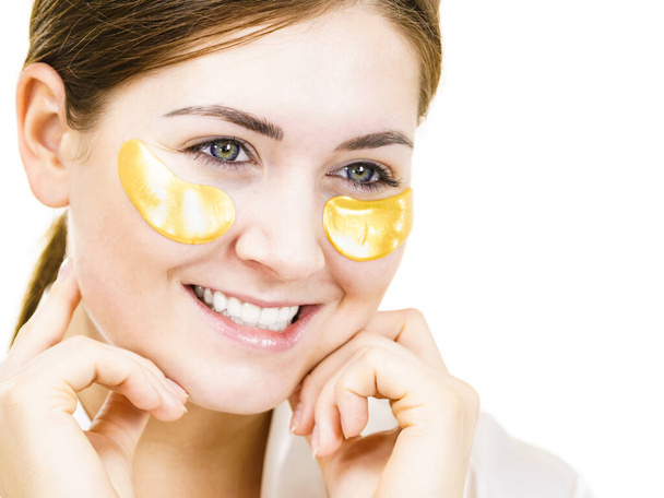 Woman applying golden collagen patches under eyes, on white. Mask removing wrinkles, dark circles. Girl taking care of delicate skin around eye. Beauty treatment. - Foto, Imagen