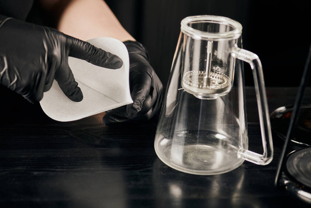 vista recortada de barista en guantes de látex negro sosteniendo filtro de papel cerca de cafetera sifón - Foto, Imagen