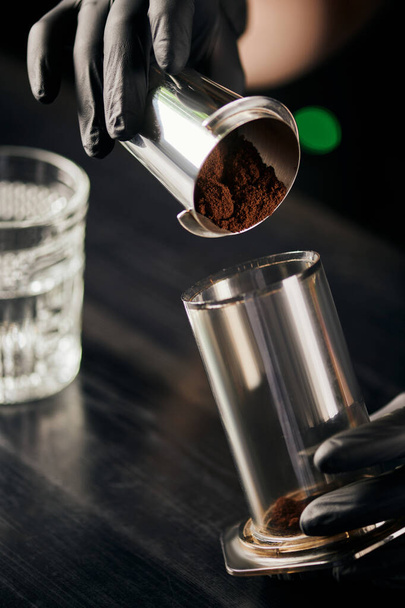 barista in black latex gloves pouring ground coffee in aero press, alternative brewing method - Foto, afbeelding