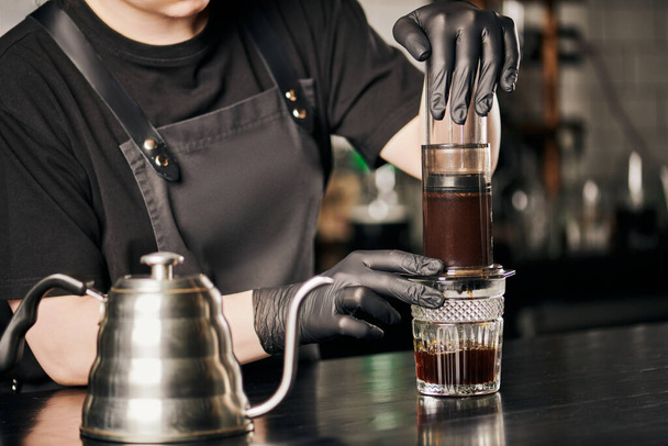 Teilansicht des Barista in schwarzen Handschuhen und Schürze bei der Zubereitung von Espresso in der Aeropresse in der Nähe des Tropfkessels - Foto, Bild