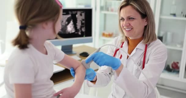 Gülümseyen doktor, liseli kızın dirsek eklemini ultrason dışbükey doğrusal dönüştürücüyle kontrol ediyor. Nazik doktor morlukları tespit etmek için ultrason yapar. - Video, Çekim