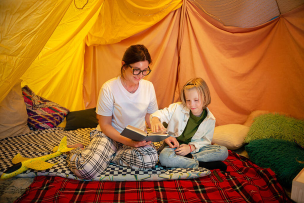 A very kind and warm looking mother is laying on her stomach next to her son reading him a book aloud and teaching him something as they are both in a comfortable large indoor tent bonding. Family - Photo, Image