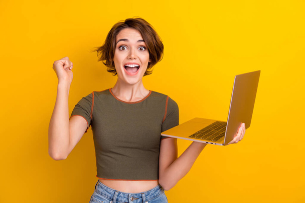 Portrait of crazy impressed woman with short hairdo wear khaki top hold laptop win gambling scream yes isolated on yellow color background. - 写真・画像