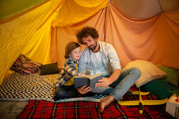 Good looking boy with long hai together with his daddy reading a story book together in the tent at home. Family - Zdjęcie, obraz