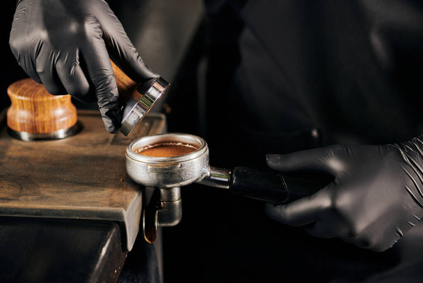 cropped view of barista in black latex gloves pressing ground coffee in portafilter, coffee shop - Zdjęcie, obraz