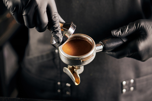 cropped view of tamper with ground coffee and portafilter in hands of barista in black gloves - Foto, immagini