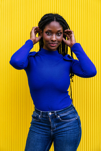Urzekający portret oszałamiającej afroamerykańskiej kobiety w żywym, kolorowym otoczeniu. Uśmiecha się dotykając włosów o żółtą ścianę. Niebieski strój dodaje uderzający kontrast. - Zdjęcie, obraz