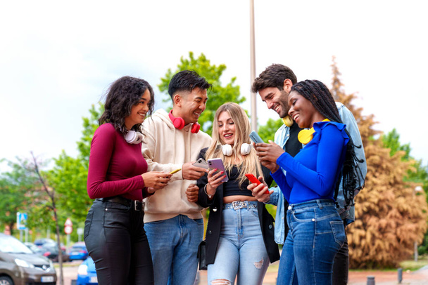 A group of five diverse young individuals, Caucasian, Chinese, and Afro-American, happily engaged with their mobile phones, wearing casual clothes and colorful headphones in a city park - Photo, Image