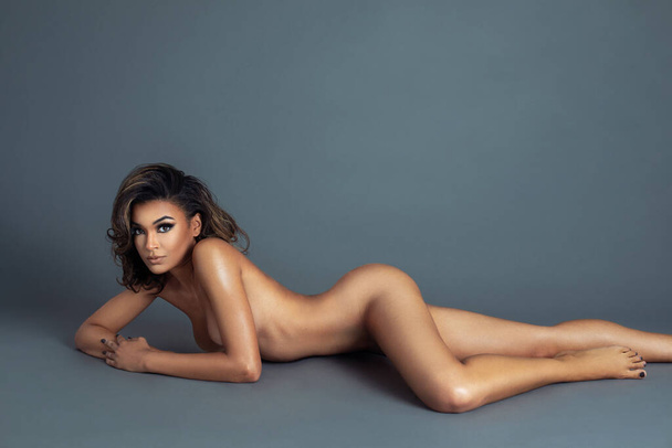 Portrait of a sensual multiracial girl with long wavy brown hair and beautiful makeup laying naked on the floor by herself inside a studio with grey background. - Foto, imagen