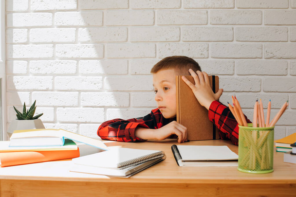 Концепция вернуться в школу. Веселый улыбающийся мальчик в процессе учебы сидит за столом с книгами, блокнотами и карандашами. Вид спереди - Фото, изображение